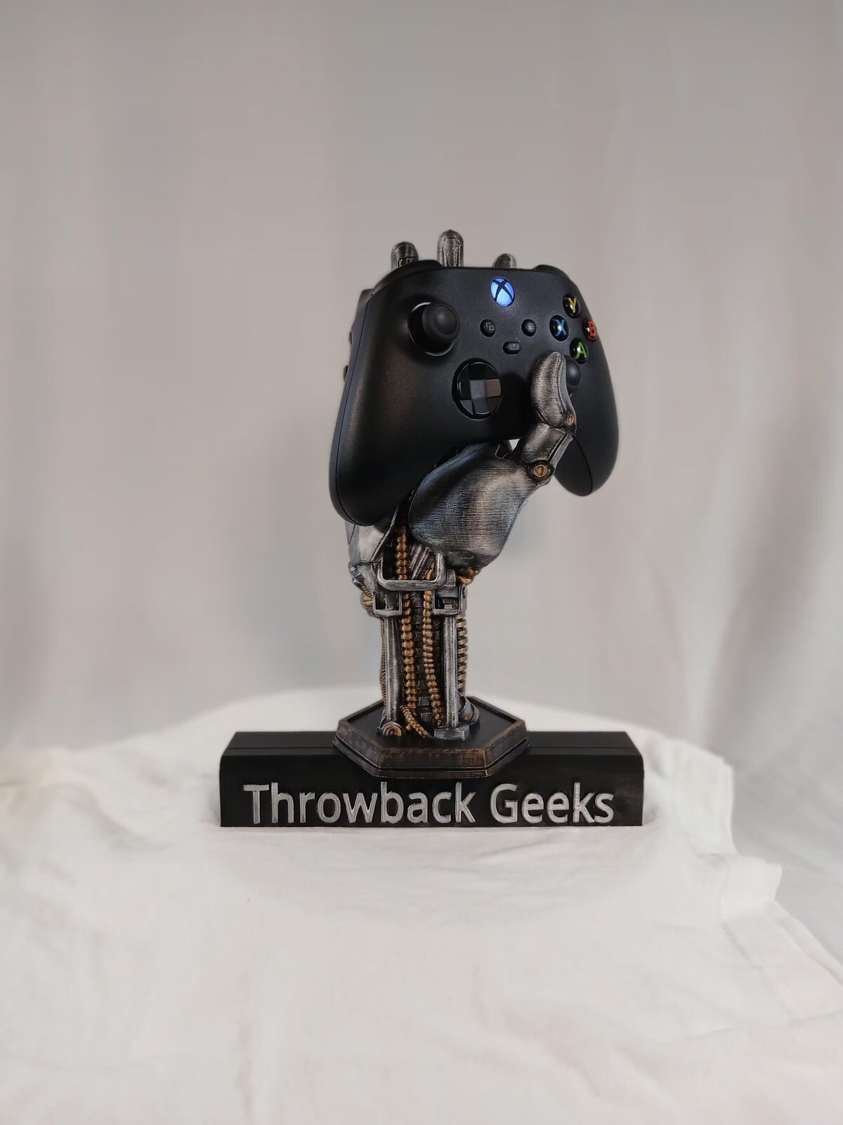 3D printed robotic hand holding an Xbox controller on a Throwback Geeks stand